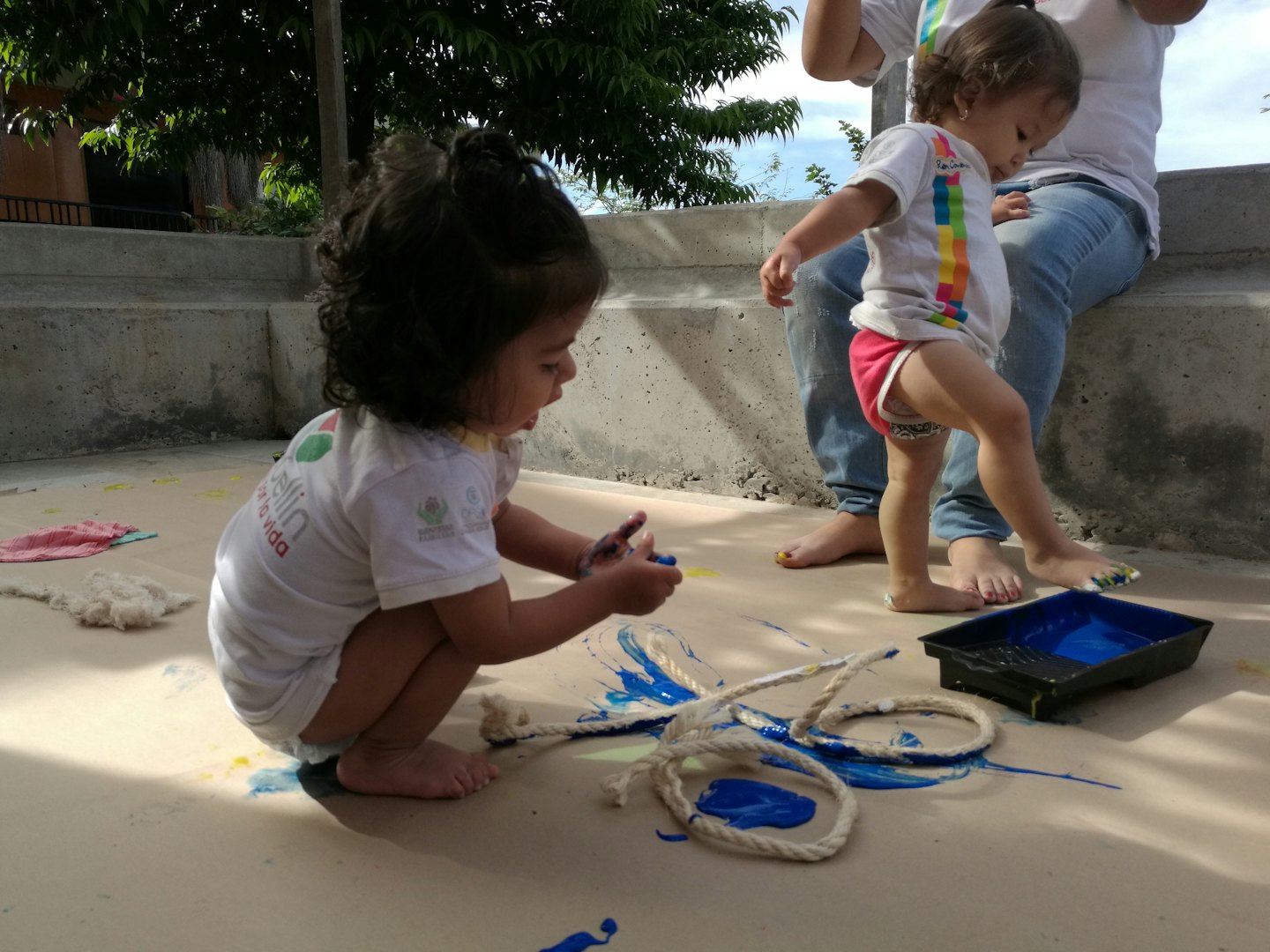 Two toddlers painting with blue paint outdoors near a tree.