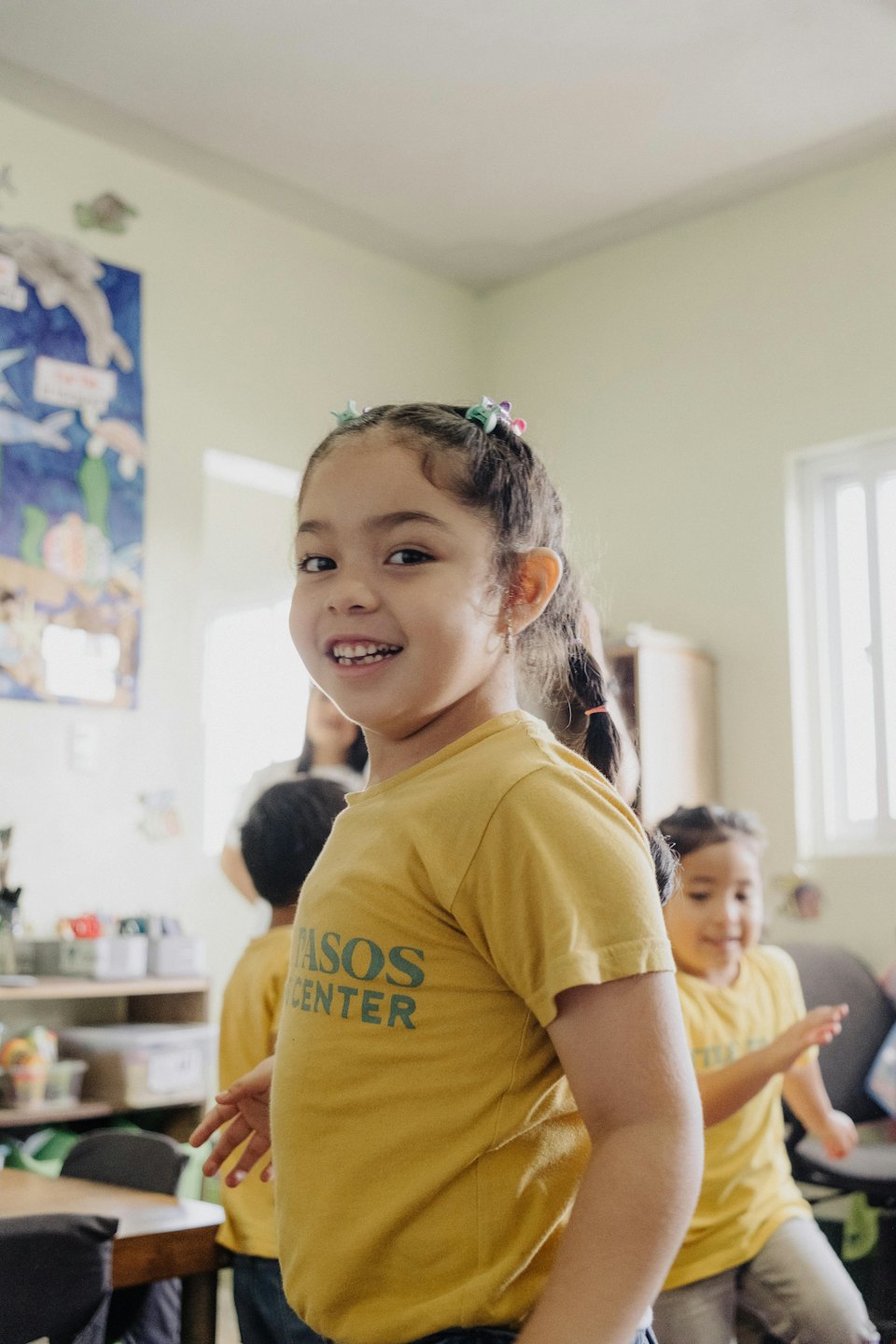 Young girl in a yellow PASOS CENTER shirt smiles in a classroom.